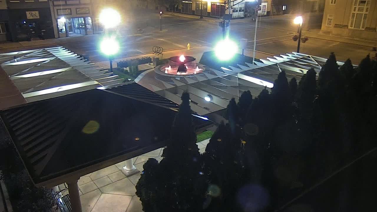 An illuminated fountain with a red flame sits in the center of a plaza at night, surrounded by dark evergreen shrubs and a modern building with a slanted roof.