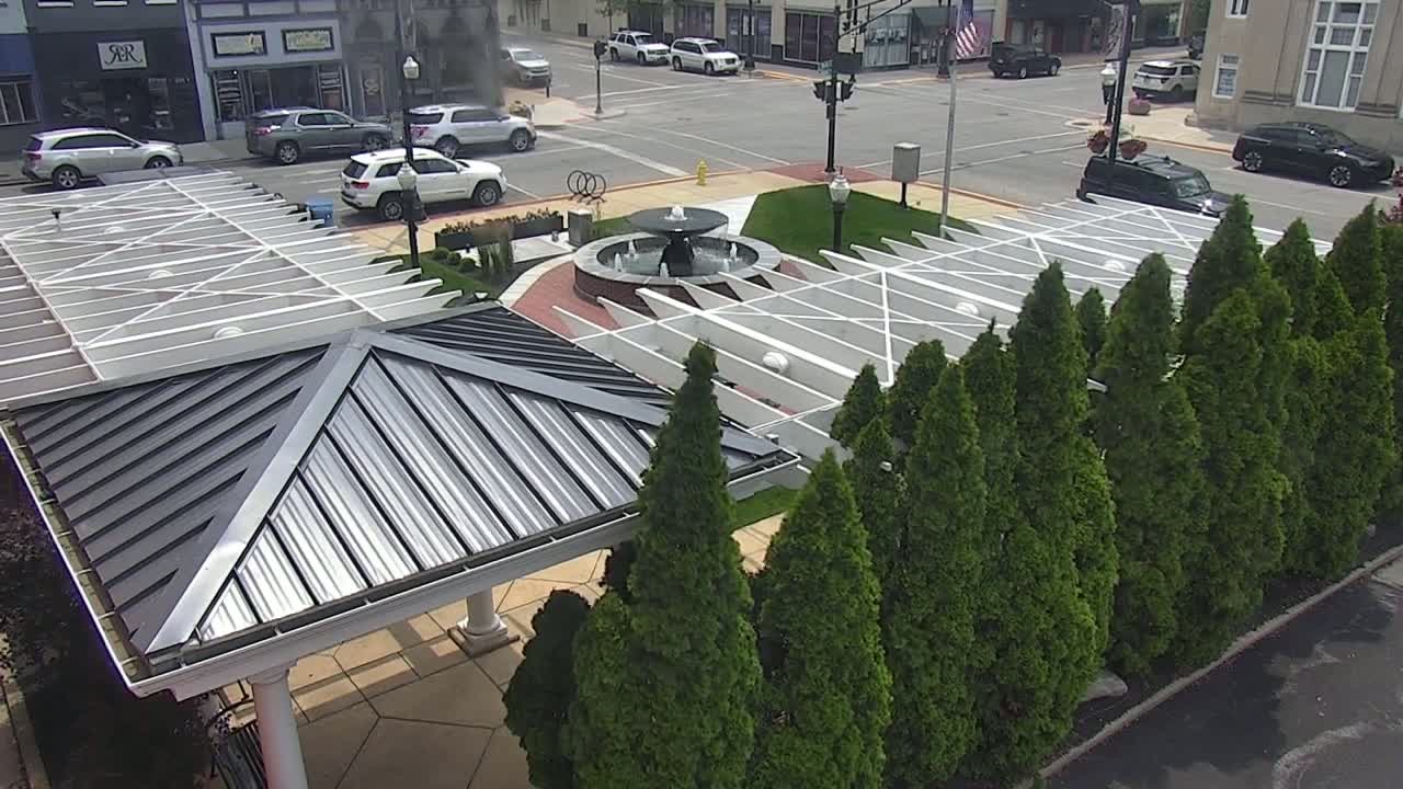 An overhead view shows a paved plaza with a central fountain, surrounded by evergreen trees and a modern pavilion with a metal roof, all under a sunny sky.