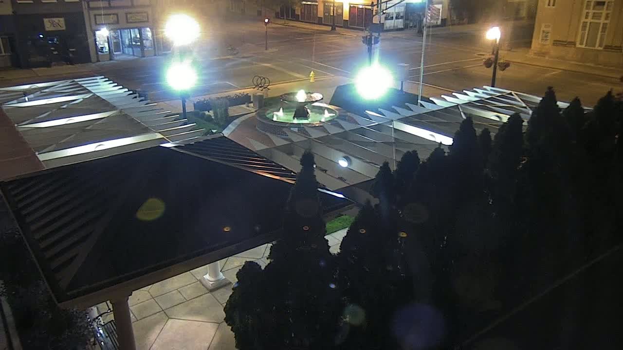 A nighttime aerial view shows a fountain in a plaza surrounded by dark evergreen trees and illuminated by streetlights.