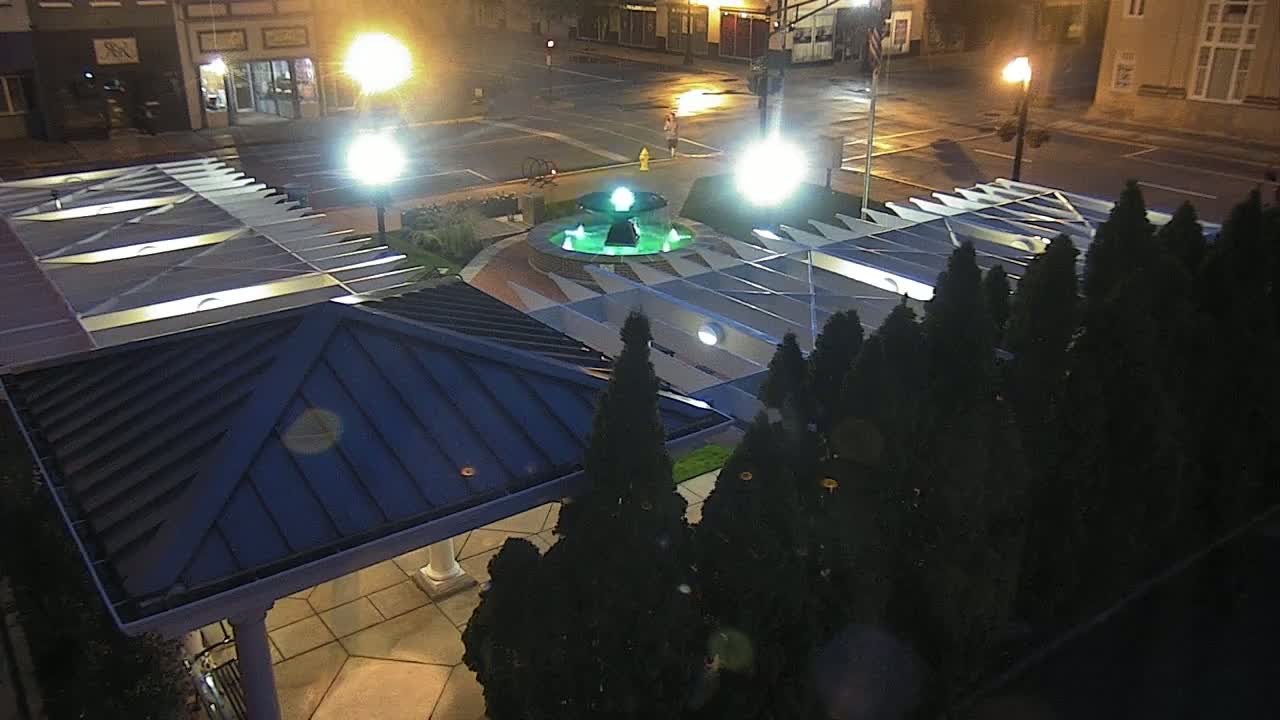 A nighttime aerial view shows a town square with a lit fountain, streetlights, and a covered pavilion surrounded by evergreen trees.