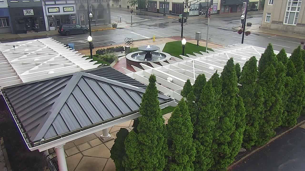 A gray-roofed gazebo sits beside a row of evergreen trees surrounding a wet, paved town square with a fountain in the center.