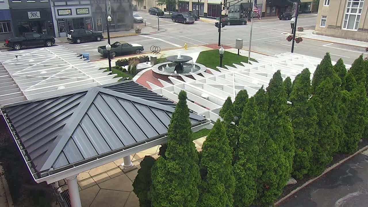 A grey-roofed gazebo sits beside a white pergola surrounding a water fountain in a town square, with evergreen trees lining the perimeter.