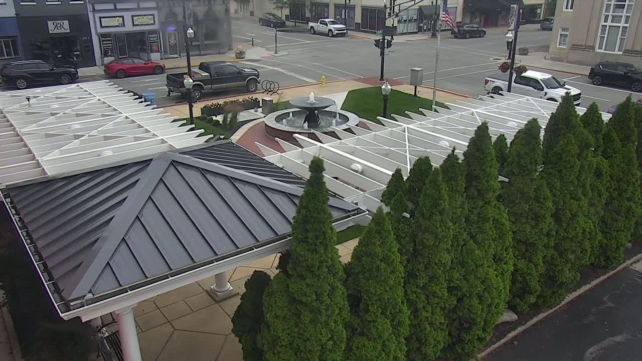 An overhead view shows a paved plaza with a water fountain, surrounded by evergreen trees and two adjacent structures with metal roofs, one white and one dark gray, under a partly cloudy sky.