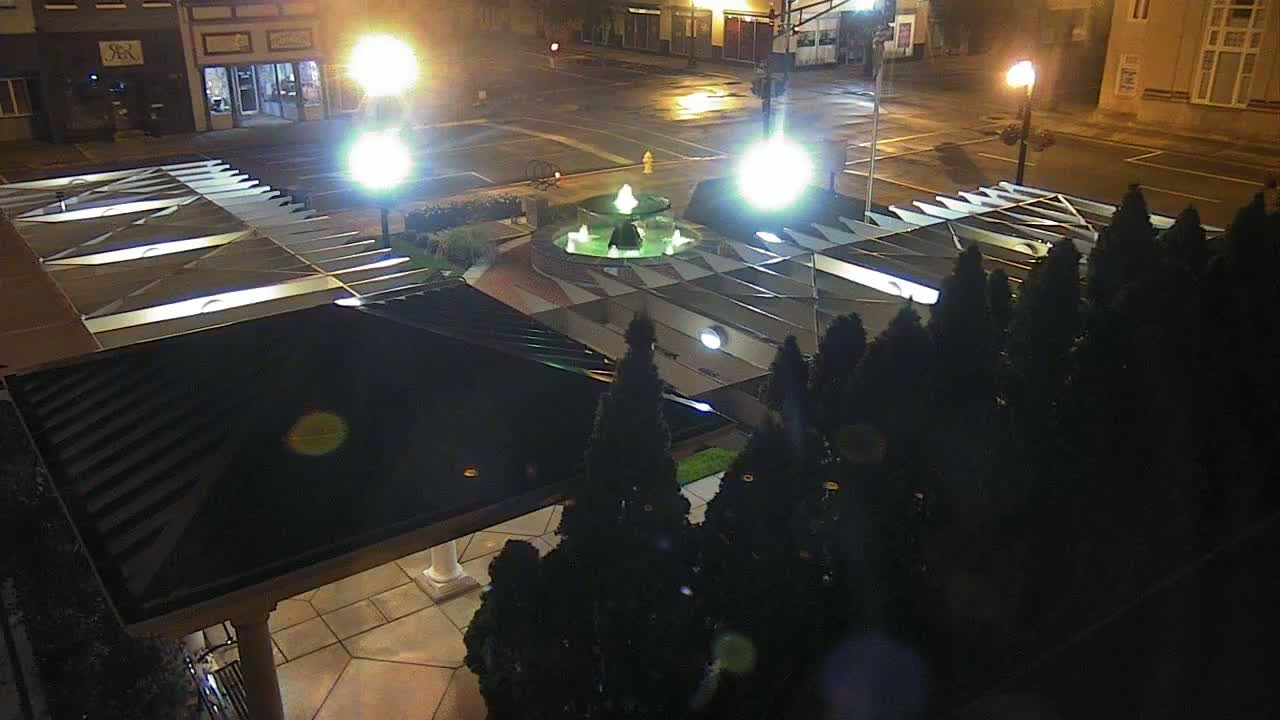 A nighttime aerial view of a town square featuring a fountain, illuminated by streetlights, surrounded by evergreen trees and a covered patio.