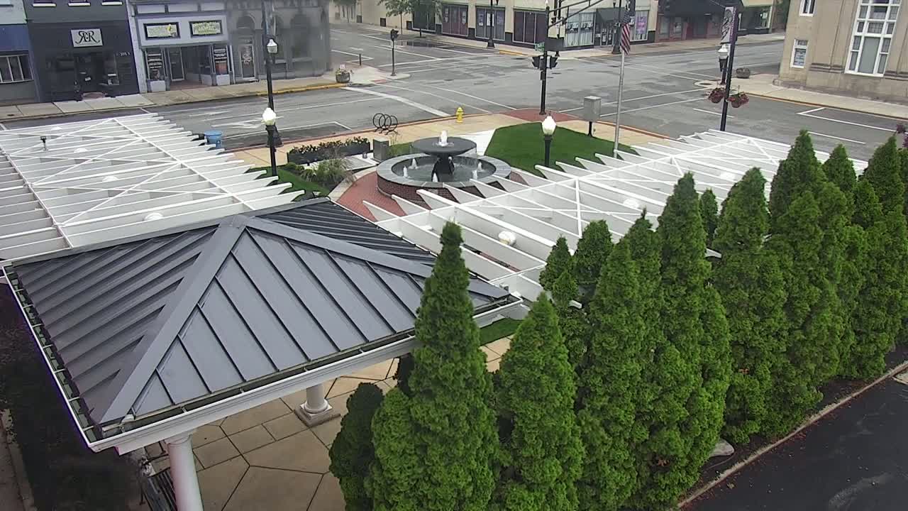 A town square with a fountain, a gray gazebo, and a row of evergreen trees under an overcast sky.