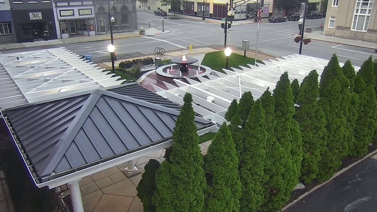 An overhead view shows a water fountain in a town square surrounded by a row of evergreen trees and a white pergola with a dark gray roof.