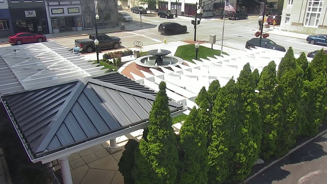 A gray-roofed pavilion with a white trellis structure surrounds a small fountain in a sunny plaza, bordered by evergreen trees and street-parked cars.