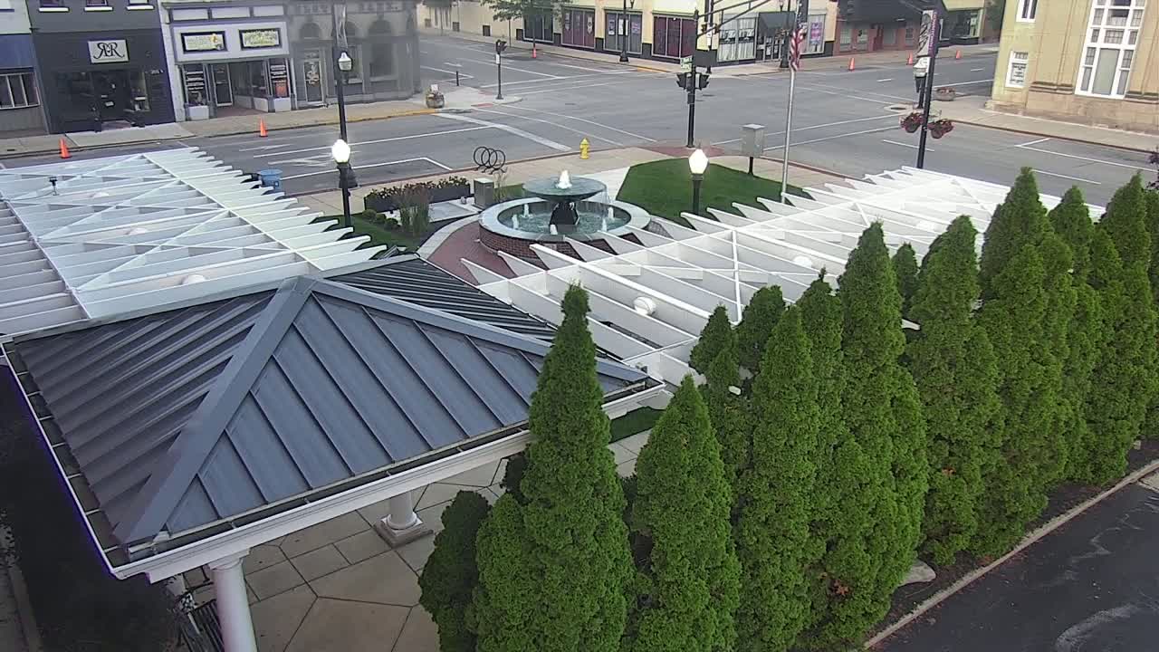An aerial view shows a gazebo with a dark gray metal roof next to a row of evergreen trees and a water fountain in a town square on a clear day.