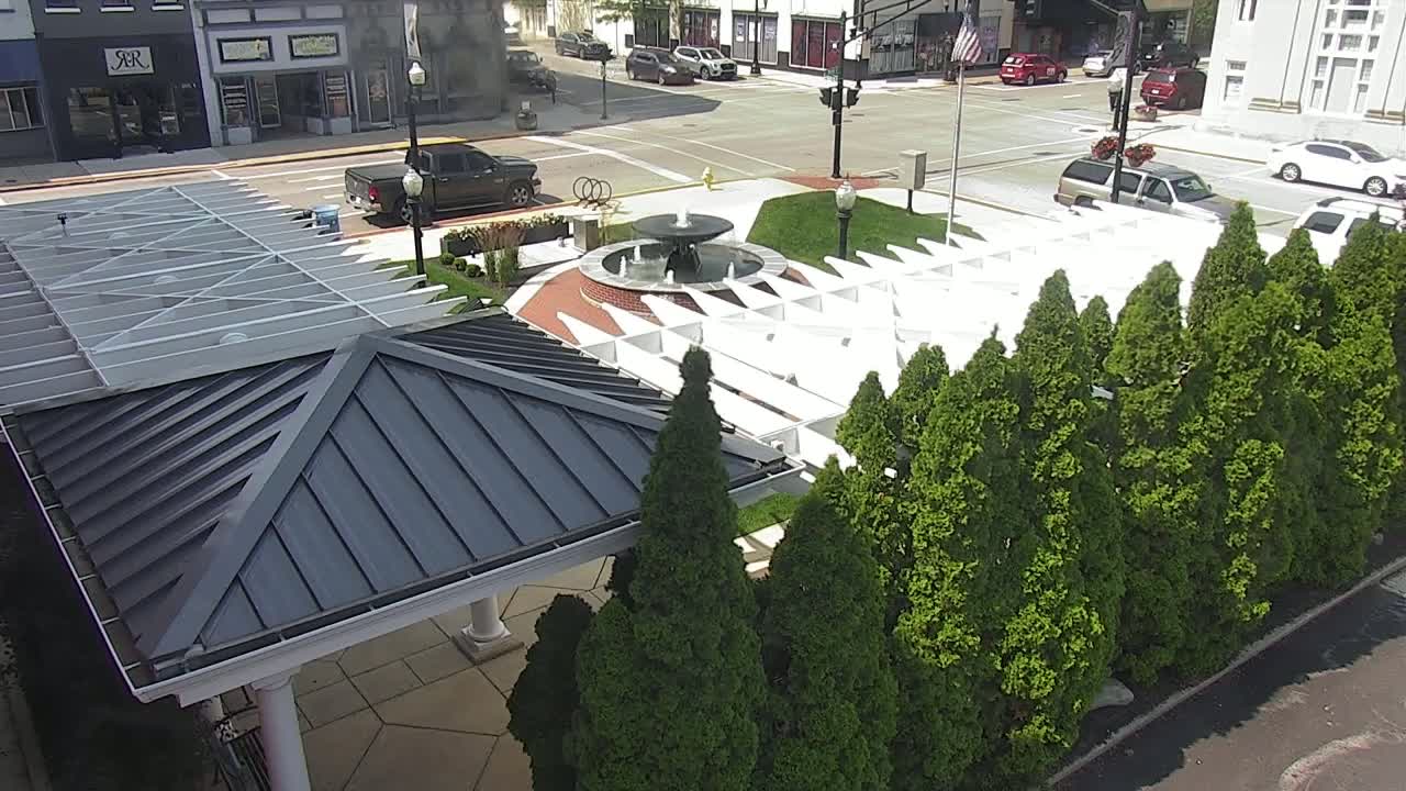 An aerial view shows a town square with a fountain, a white pergola, and a row of evergreen trees on a sunny day.