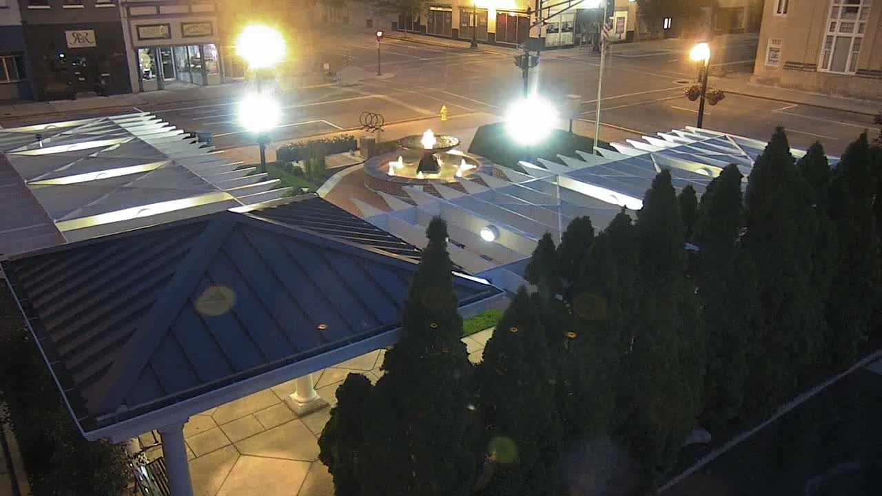 An overhead nighttime view of a town square featuring a fountain, a gazebo, and neatly trimmed evergreen trees under bright streetlights.