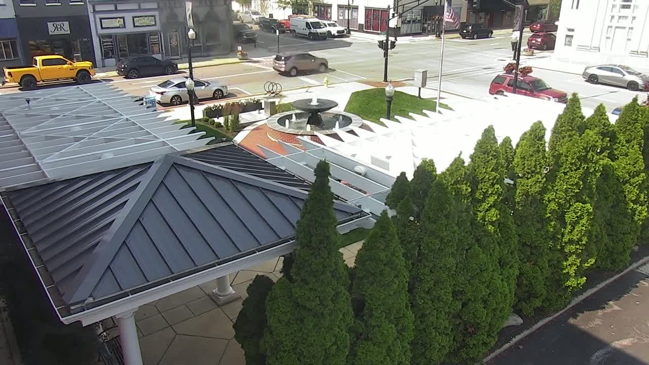 A dark-gray roofed gazebo sits beside a row of evergreen trees next to a town square fountain on a sunny day.