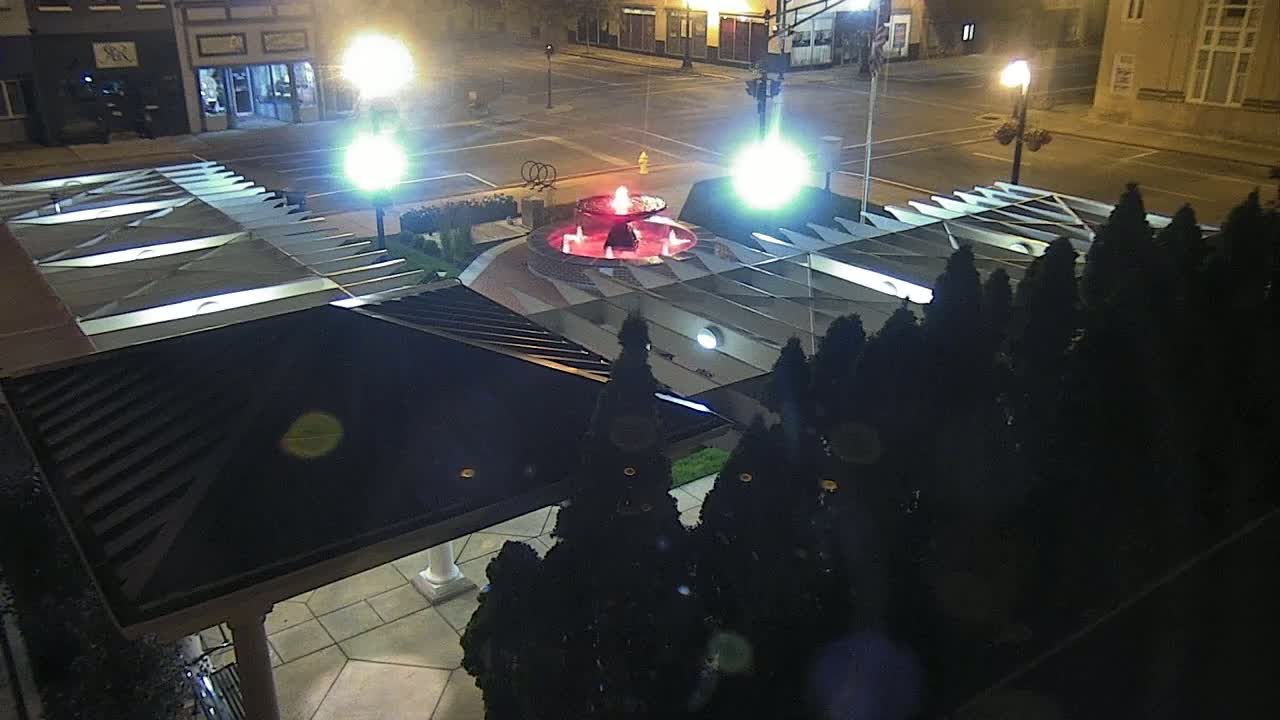 An illuminated fountain is at the center of a plaza at night, surrounded by dark evergreen shrubs and streetlights, viewed from above a dark-roofed structure.