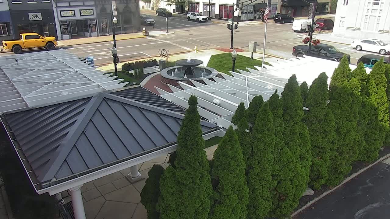 An aerial view shows a town square with a fountain, a gazebo, and a row of evergreen trees on a sunny day.