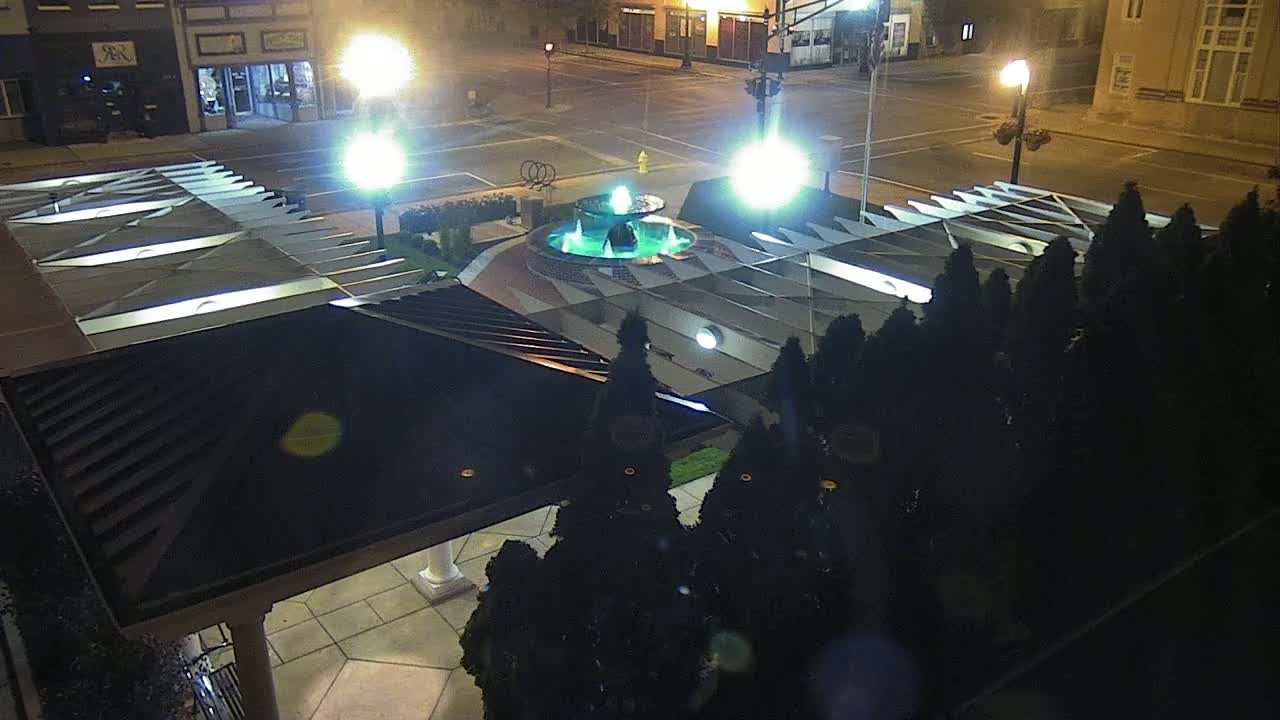 A nighttime view of a fountain surrounded by dark evergreen shrubs and illuminated by bright lights, situated in a town square.