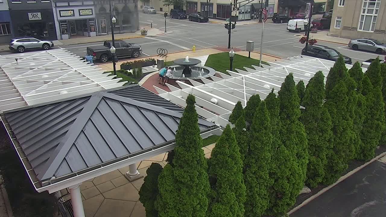 An overhead view shows a gazebo with a dark gray metal roof next to a row of evergreen trees surrounding a central fountain in a town square on a partly cloudy day.