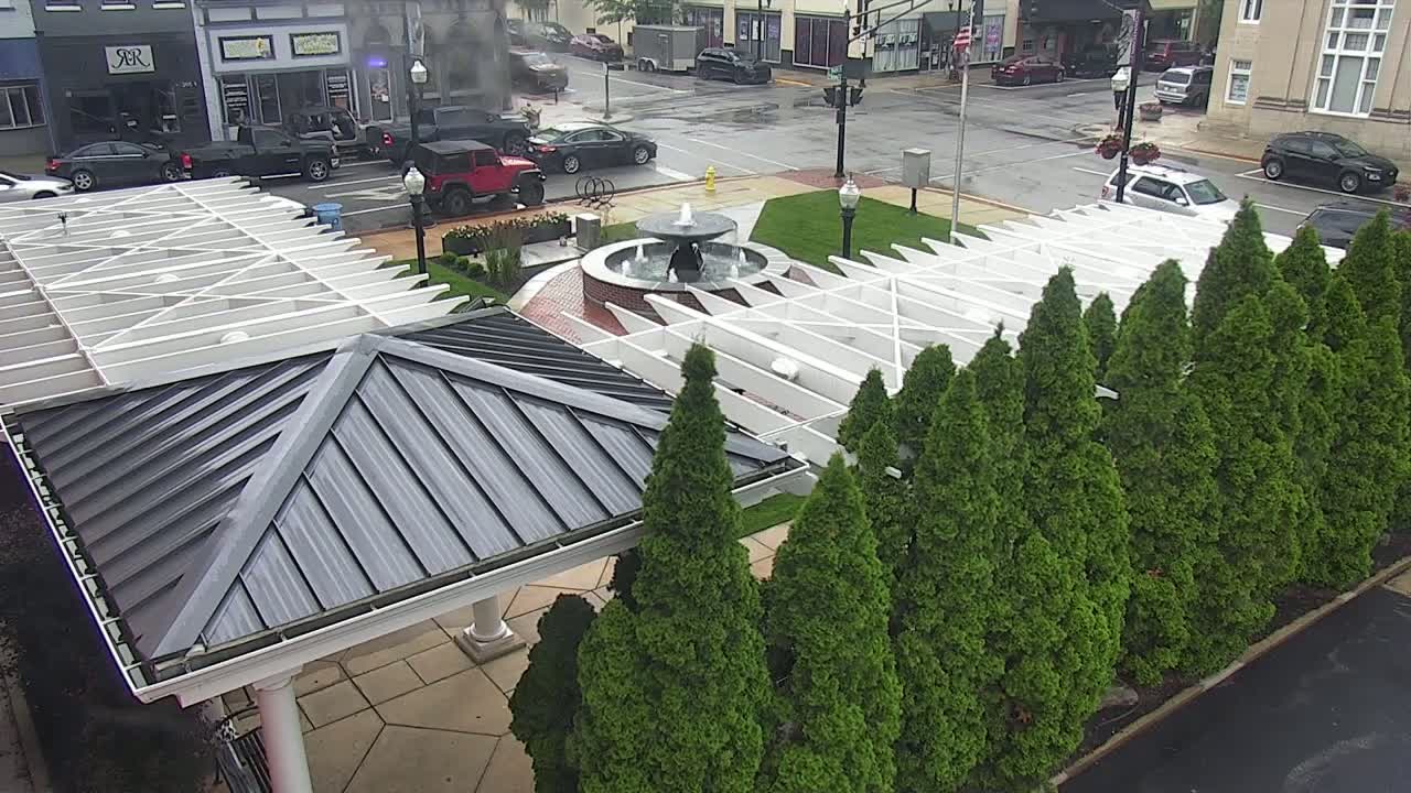 A wet town square features a central fountain, surrounded by evergreen trees and white pergola structures, under a mostly cloudy sky.