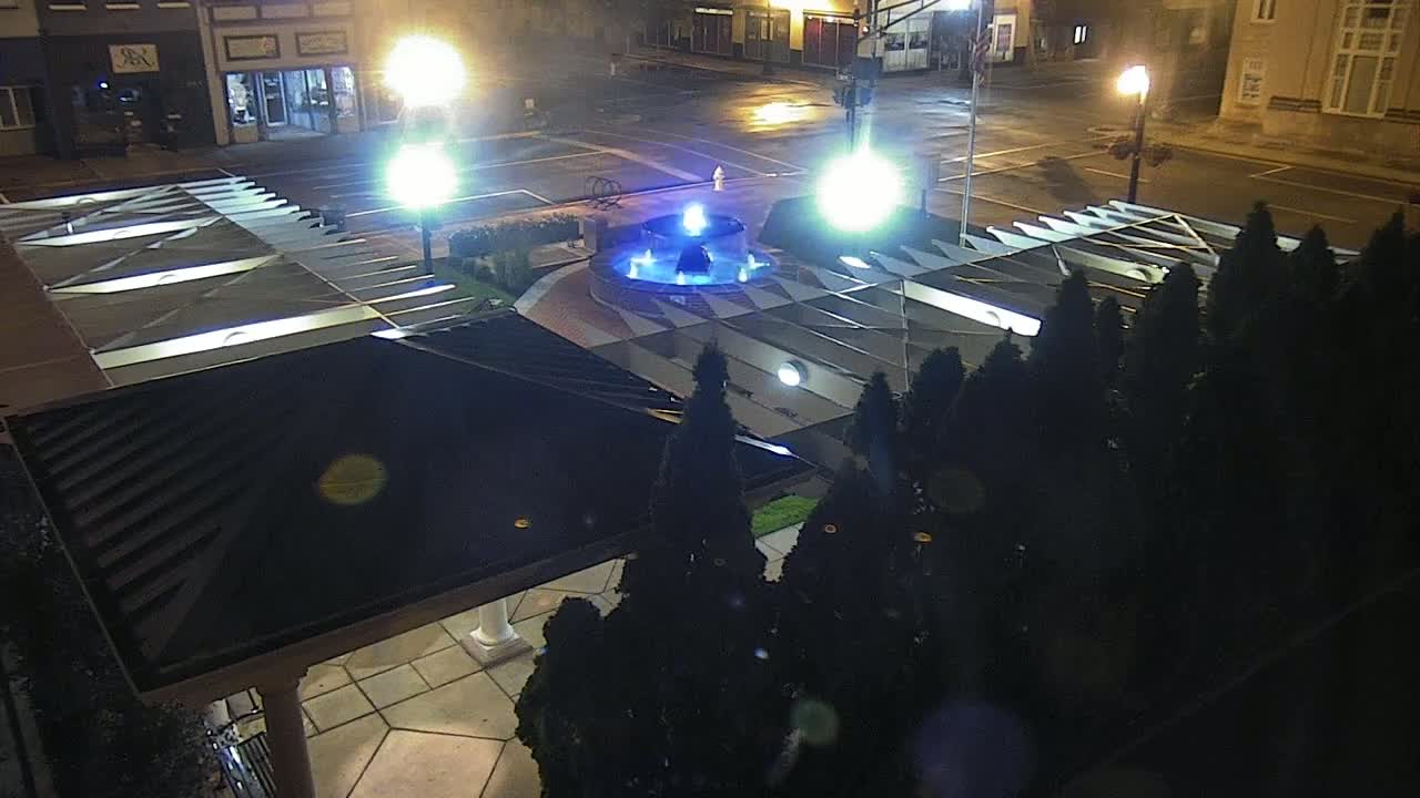 A nighttime aerial view shows a blue-lit fountain in a plaza surrounded by evergreen shrubs and illuminated by streetlights.