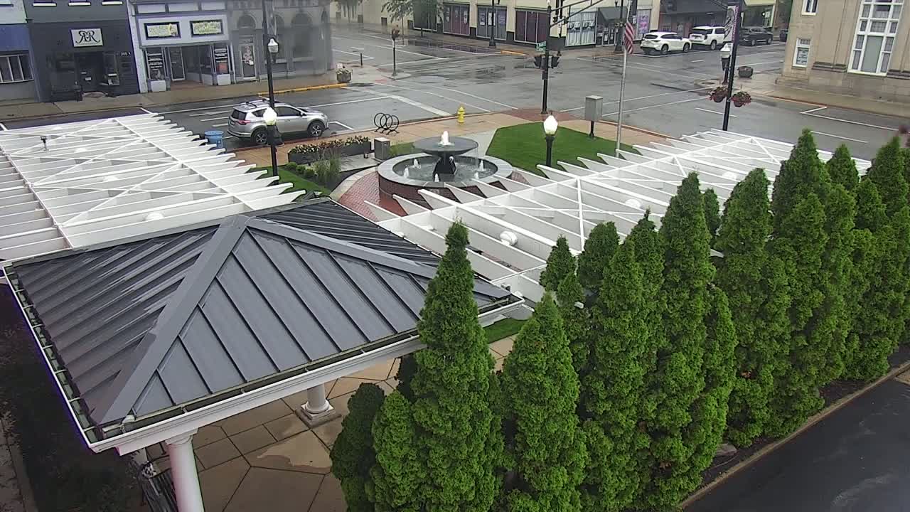 A wet, paved town square features a fountain surrounded by evergreen trees and two partially visible pavilions with white and dark gray roofs.