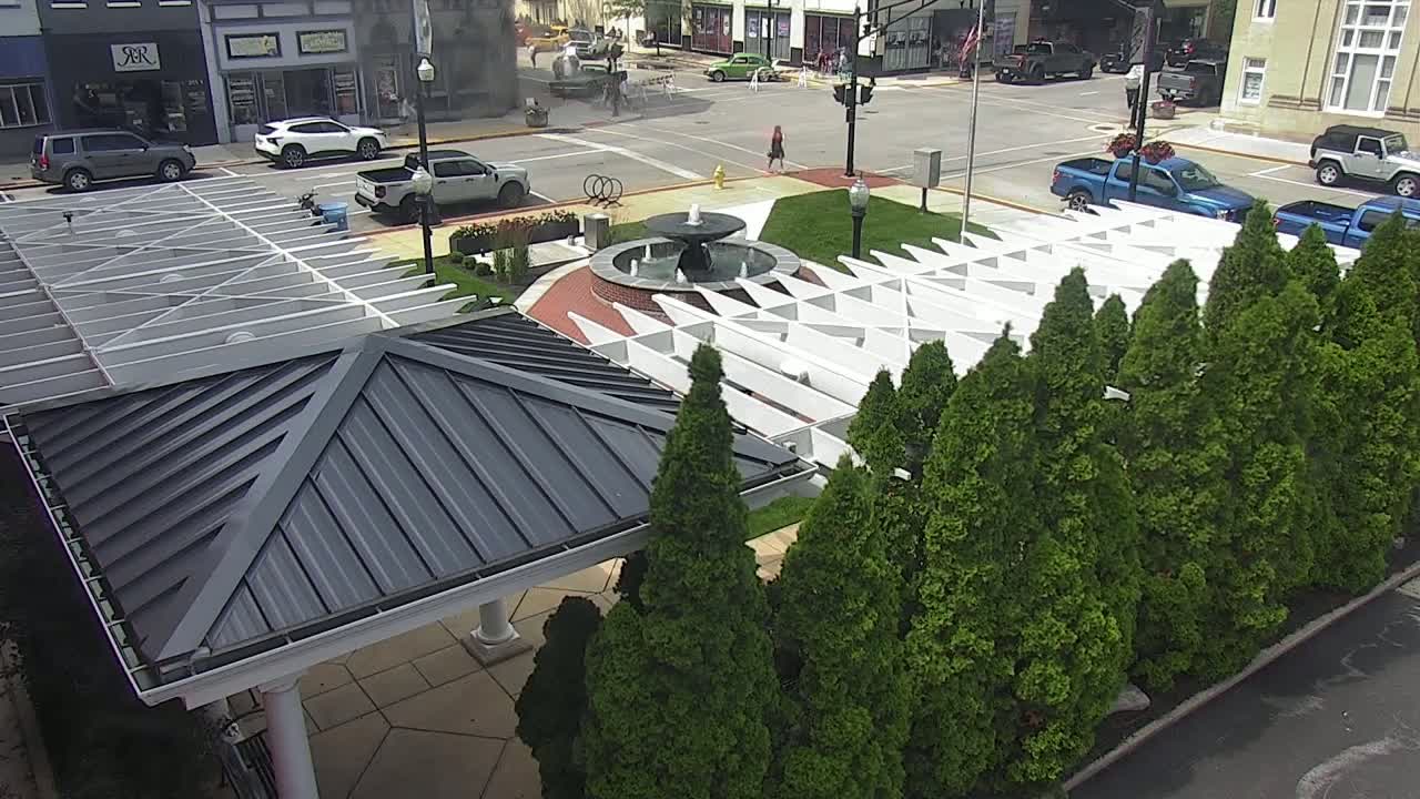 An aerial view shows a town square with a fountain, parked cars, and a white pergola structure partially obscured by a row of evergreen trees on a sunny day.