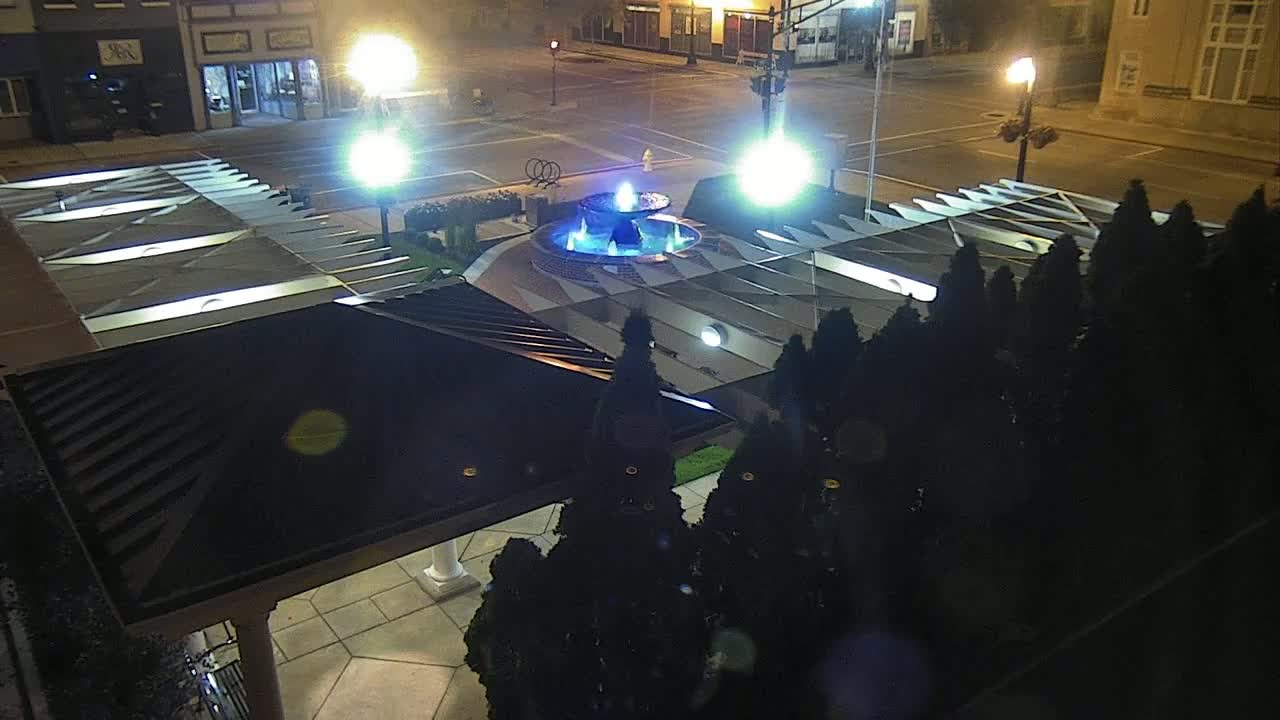 A nighttime aerial view of a plaza with a lit fountain surrounded by dark evergreen shrubs and a portion of a building's roof.