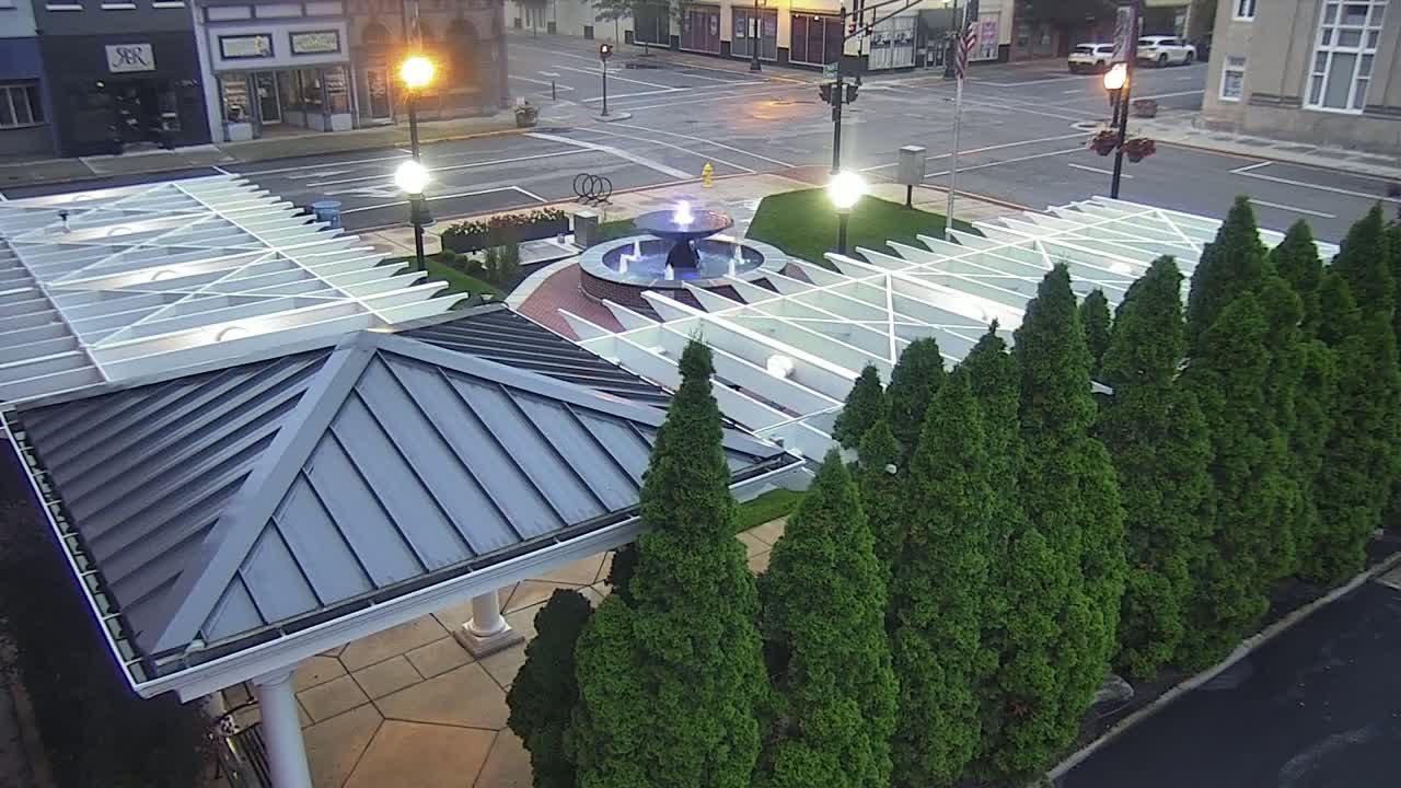 An aerial view shows a town square at dusk with a water fountain surrounded by a white pergola and a row of evergreen trees.