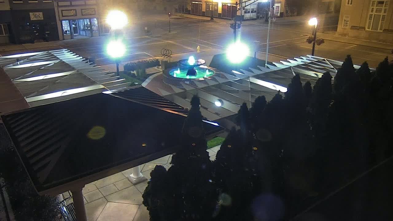 An illuminated plaza at night features a central fountain surrounded by dark evergreen shrubs and a modern, angled roof structure.