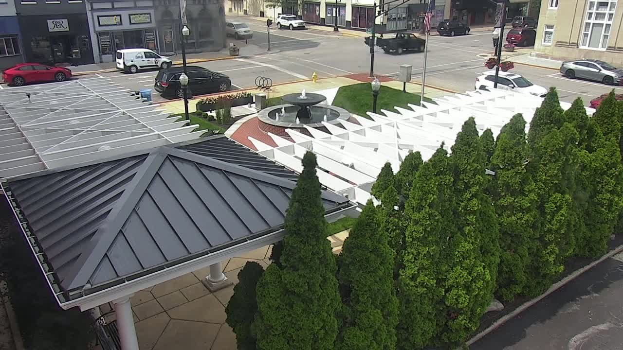 An overhead view shows a town square with a fountain, a white pergola structure, and a row of evergreen trees, on a sunny day.