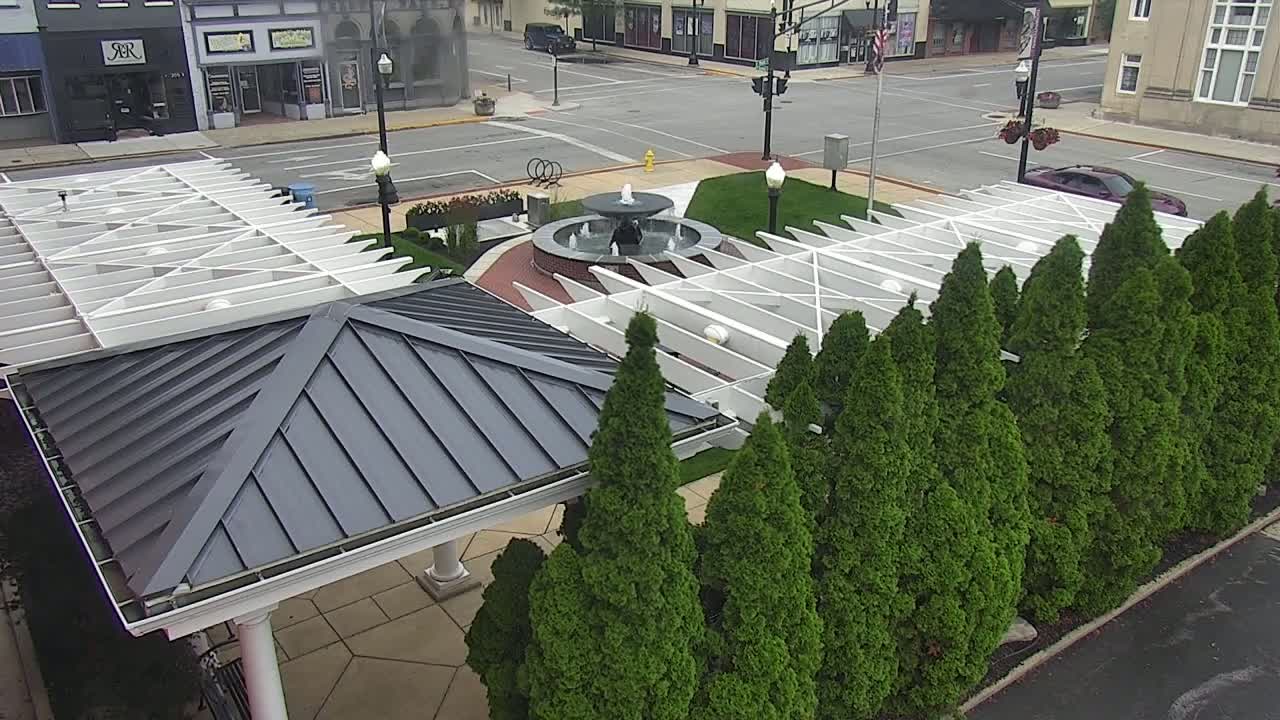 A dark-gray roofed gazebo with a white pergola sits beside a row of evergreen trees surrounding a circular fountain in a town square on a partly cloudy day.