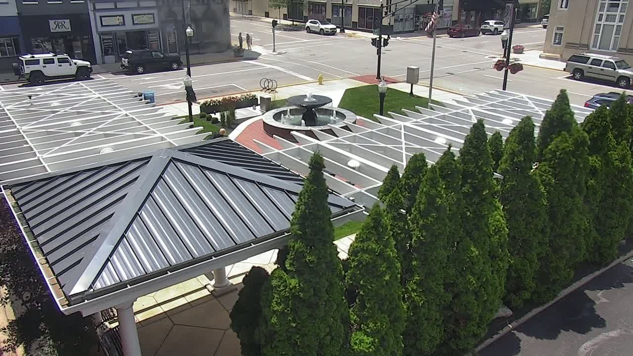 An aerial view shows a plaza with a central fountain, surrounded by a row of evergreen trees and modern pavilions under a sunny sky.