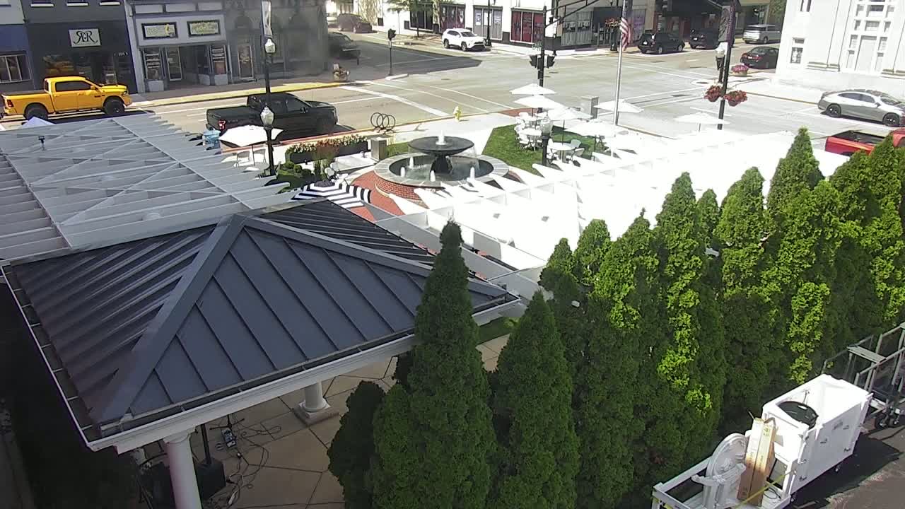 A courtyard with a fountain, white lounge chairs, and numerous green trees under a partly sunny sky, is adjacent to a dark-grey roofed gazebo and a street with parked cars.