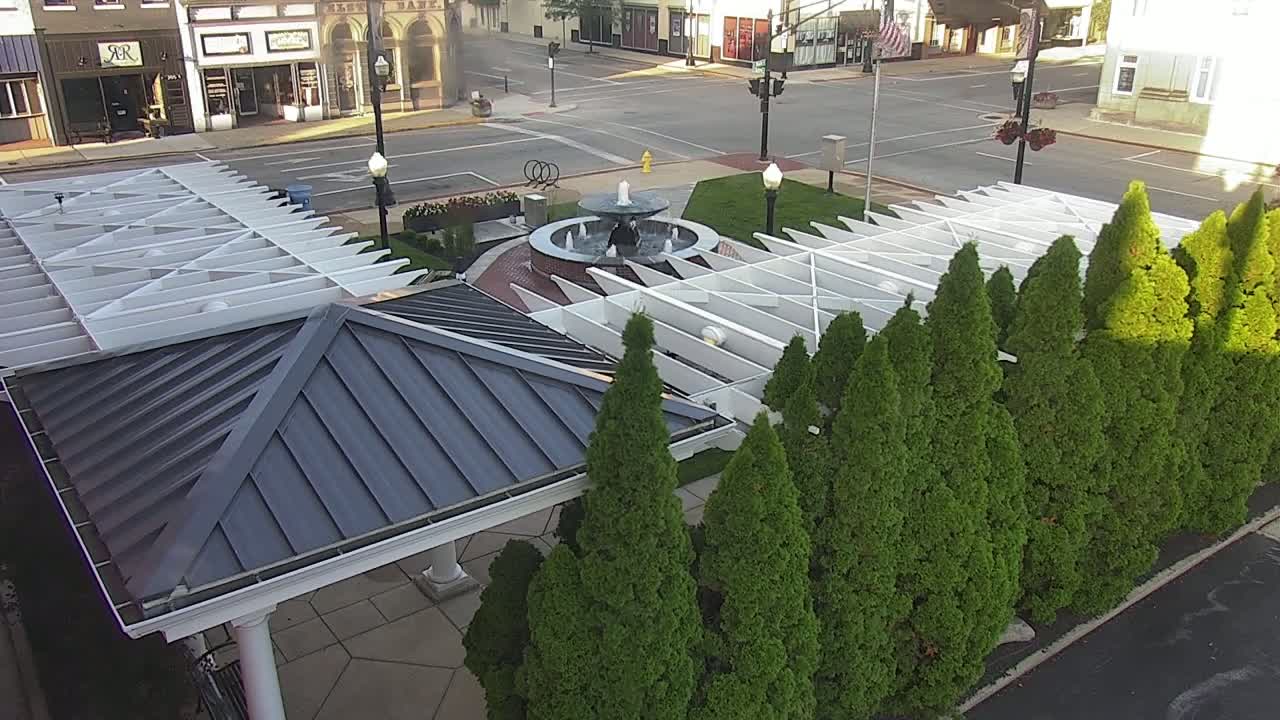 An aerial view shows a gazebo with a dark gray metal roof next to a row of evergreen trees surrounding a central fountain in a town square on a sunny day.