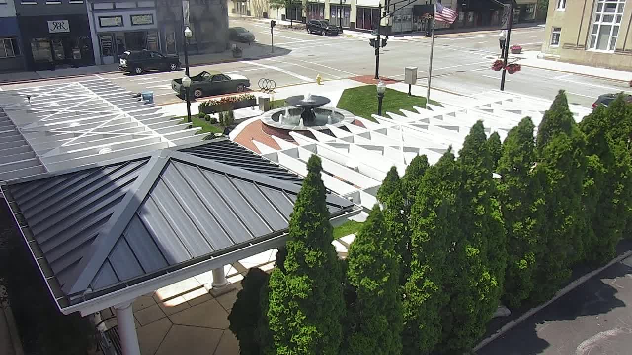 An overhead view of a plaza featuring a fountain, a white geometric structure, and a row of evergreen trees on a sunny day.
