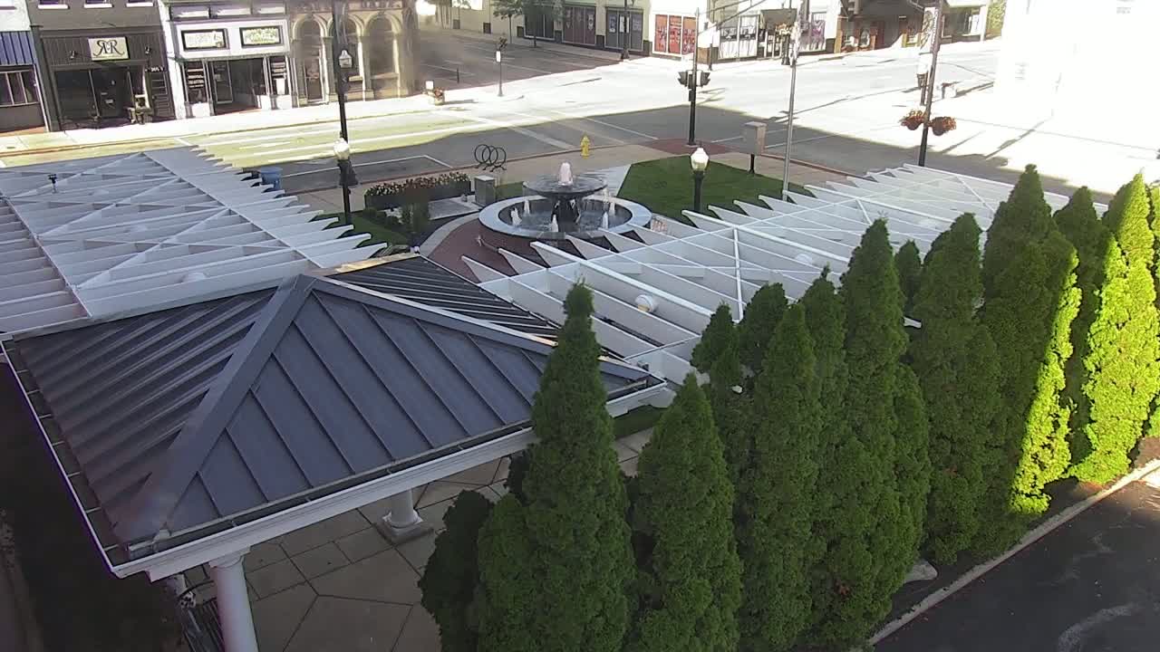 A paved plaza with a central fountain is partially shaded by a white pergola and surrounded by evergreen trees on a sunny day.