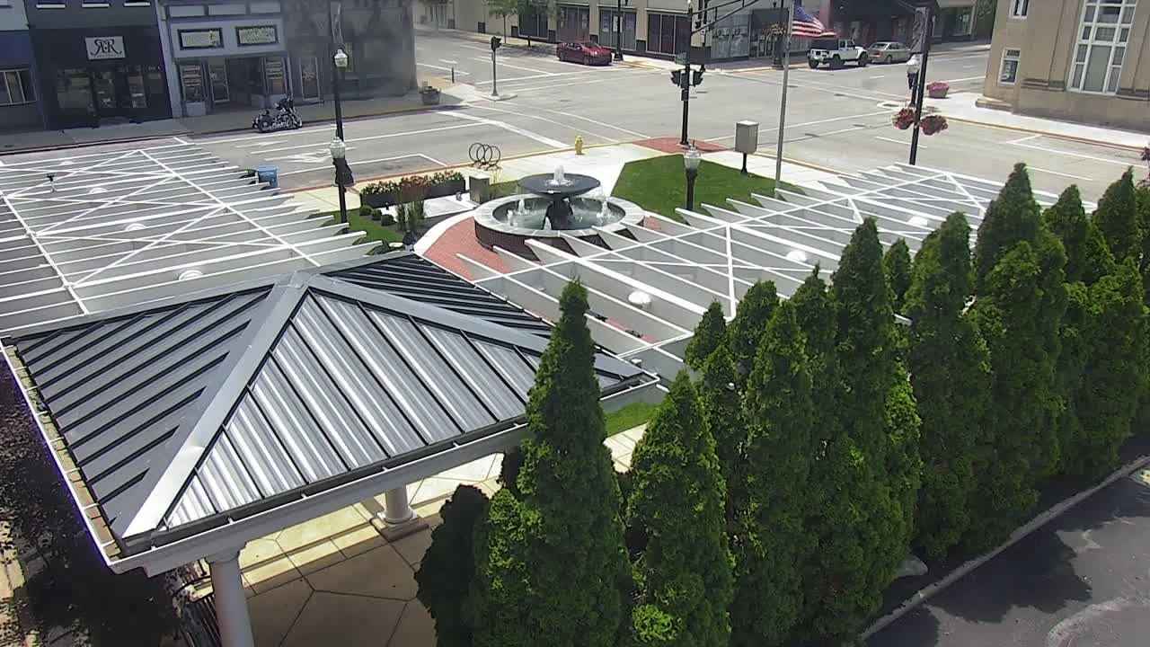 An aerial view shows a town square with a central fountain, surrounded by a geometric pattern of paving stones and rows of evergreen trees, all under a sunny sky.