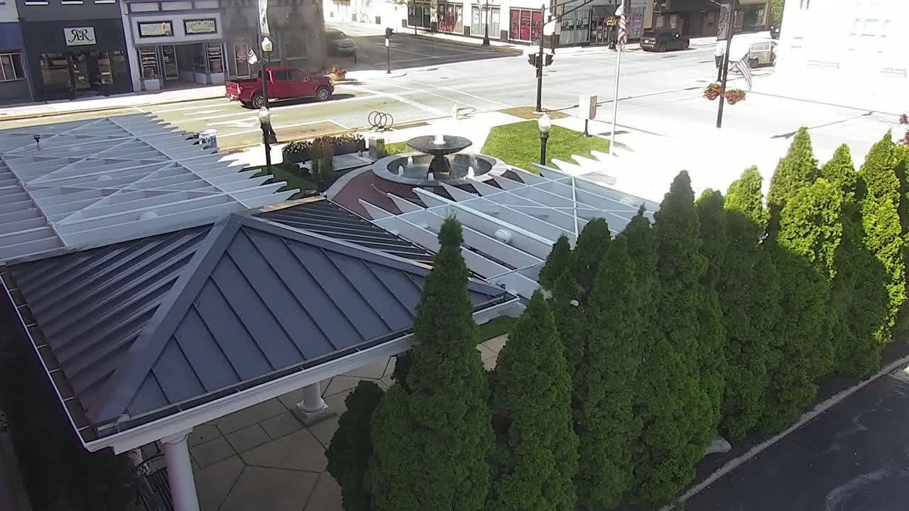 A dark gray metal roofed gazebo with a white pergola sits adjacent to a fountain in a landscaped area with a row of evergreen trees, on a sunny day.