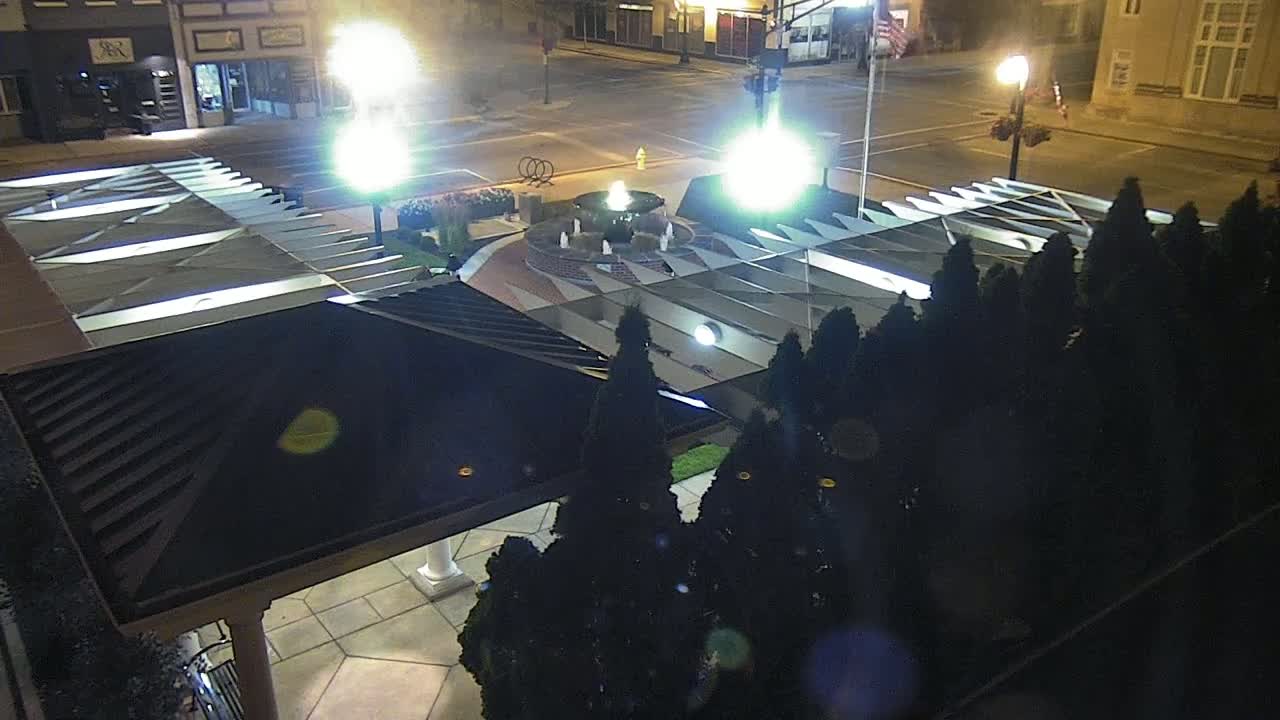 A nighttime, overhead view of a town square featuring a fountain illuminated by bright lights, surrounded by dark evergreen shrubs and a covered walkway.