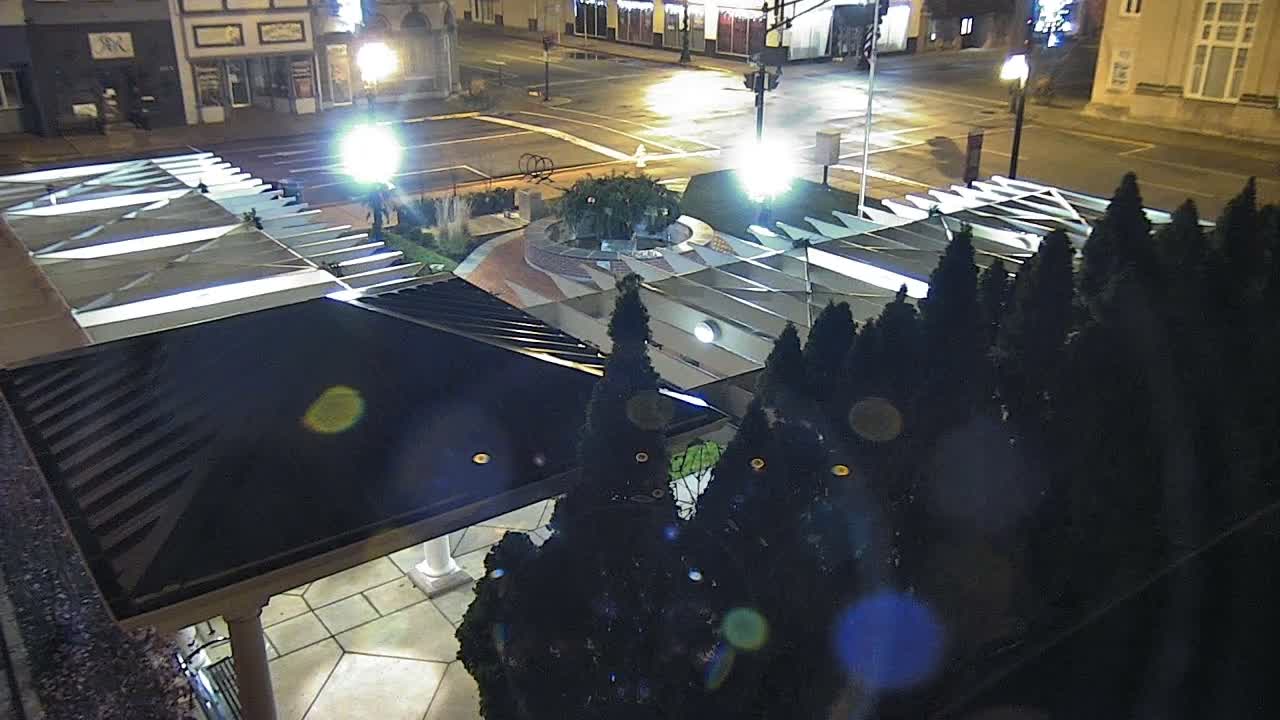 An elevated nighttime view reveals a public plaza featuring a large segmented white canopy and dark evergreen trees, with wet streets reflecting bright streetlights under damp conditions.
