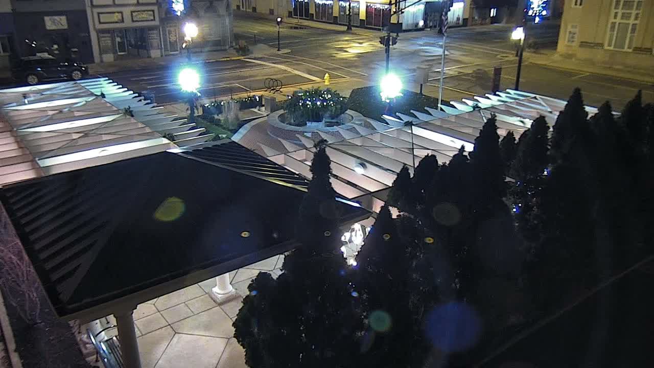 An elevated nighttime view shows a brightly lit urban plaza featuring a geometric canopy structure and a circular planter with decorative lights, adjacent to a reflective street lined with buildings, all under clear weather conditions.