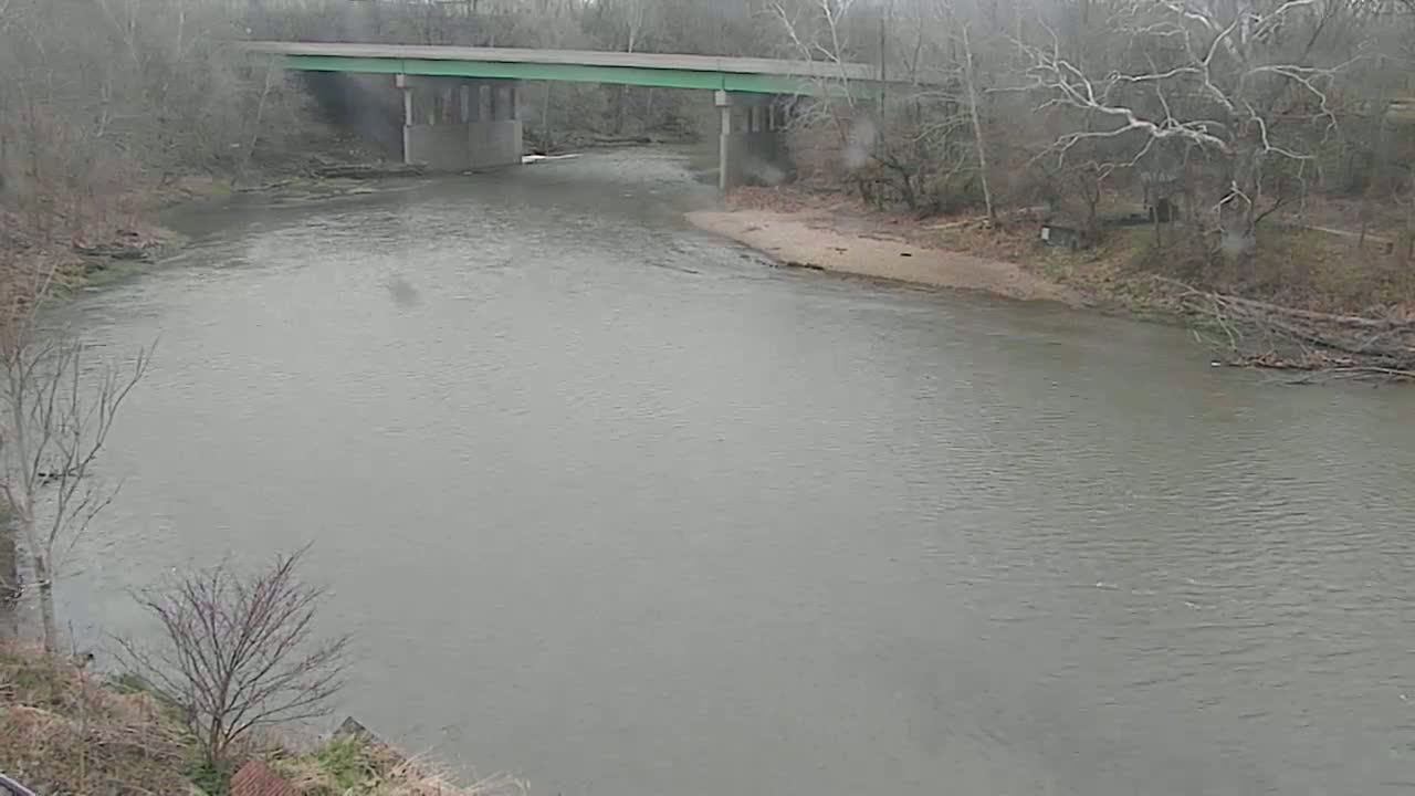 A muted green bridge spans a wide, calm river under an overcast sky, surrounded by bare winter trees and a sandy riverbank.