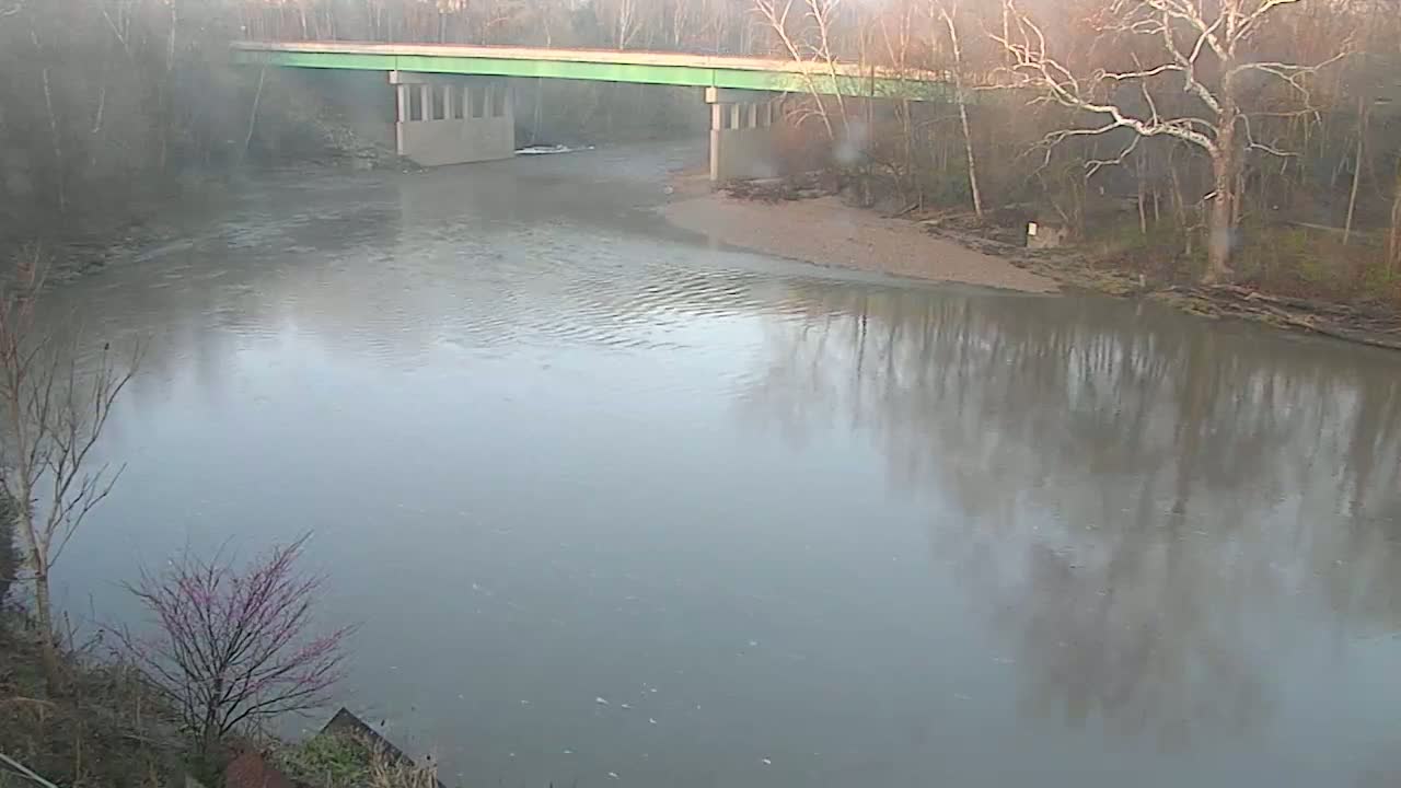 A wide, calm river reflects the bare winter trees and a green-topped bridge on an overcast day.