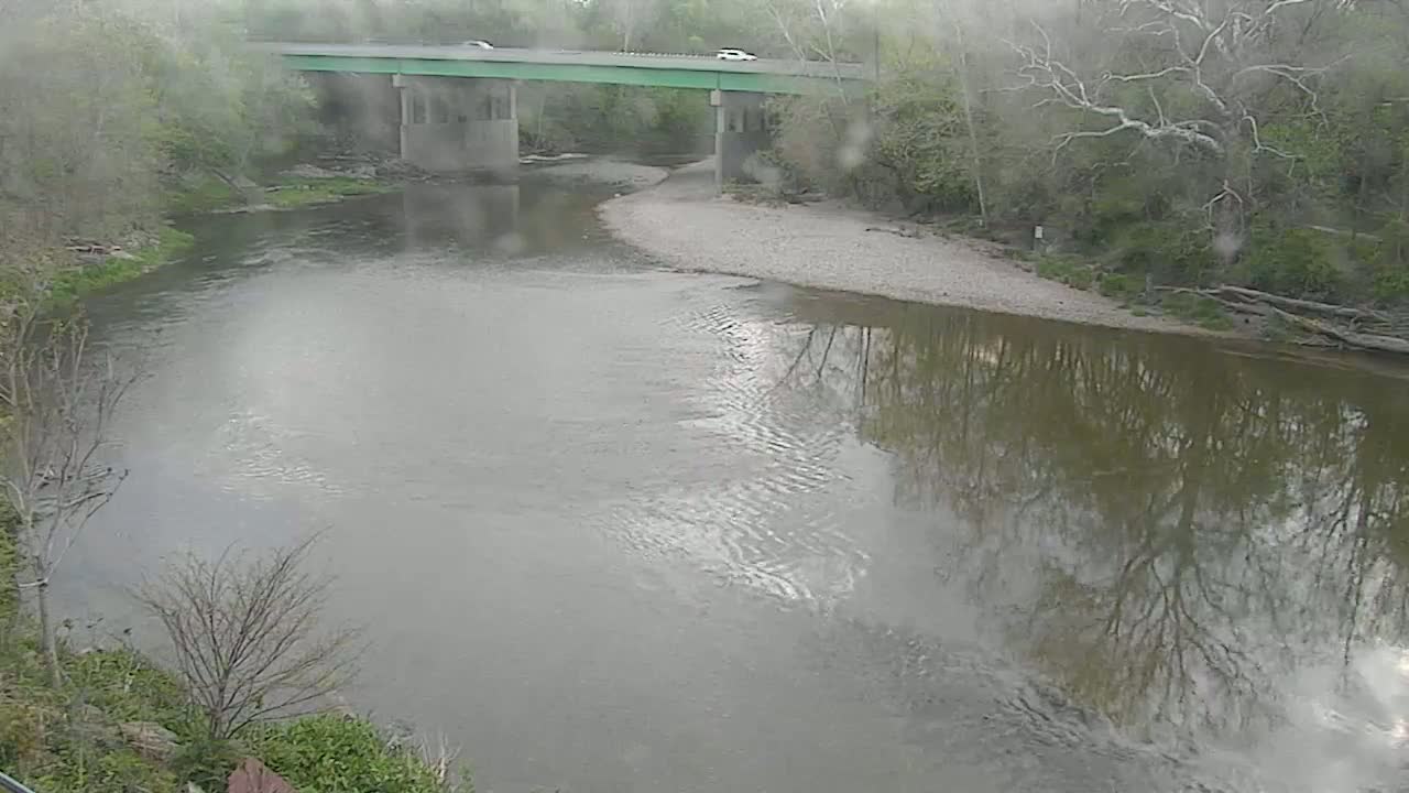 A river flows under a bridge on a rainy day, reflecting the surrounding trees and overcast sky.