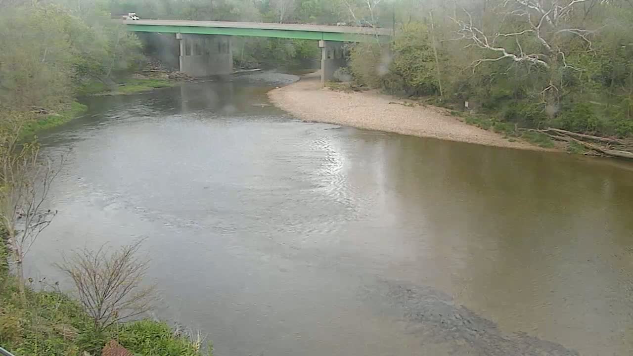 A river bends beneath a green bridge on a rainy day, surrounded by trees and a muddy riverbank.
