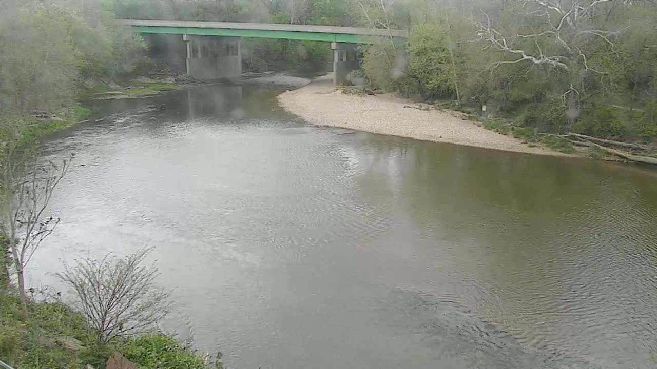 A gray-brown river flows under a green bridge on a cloudy day, with trees and a gravel riverbank visible on either side.