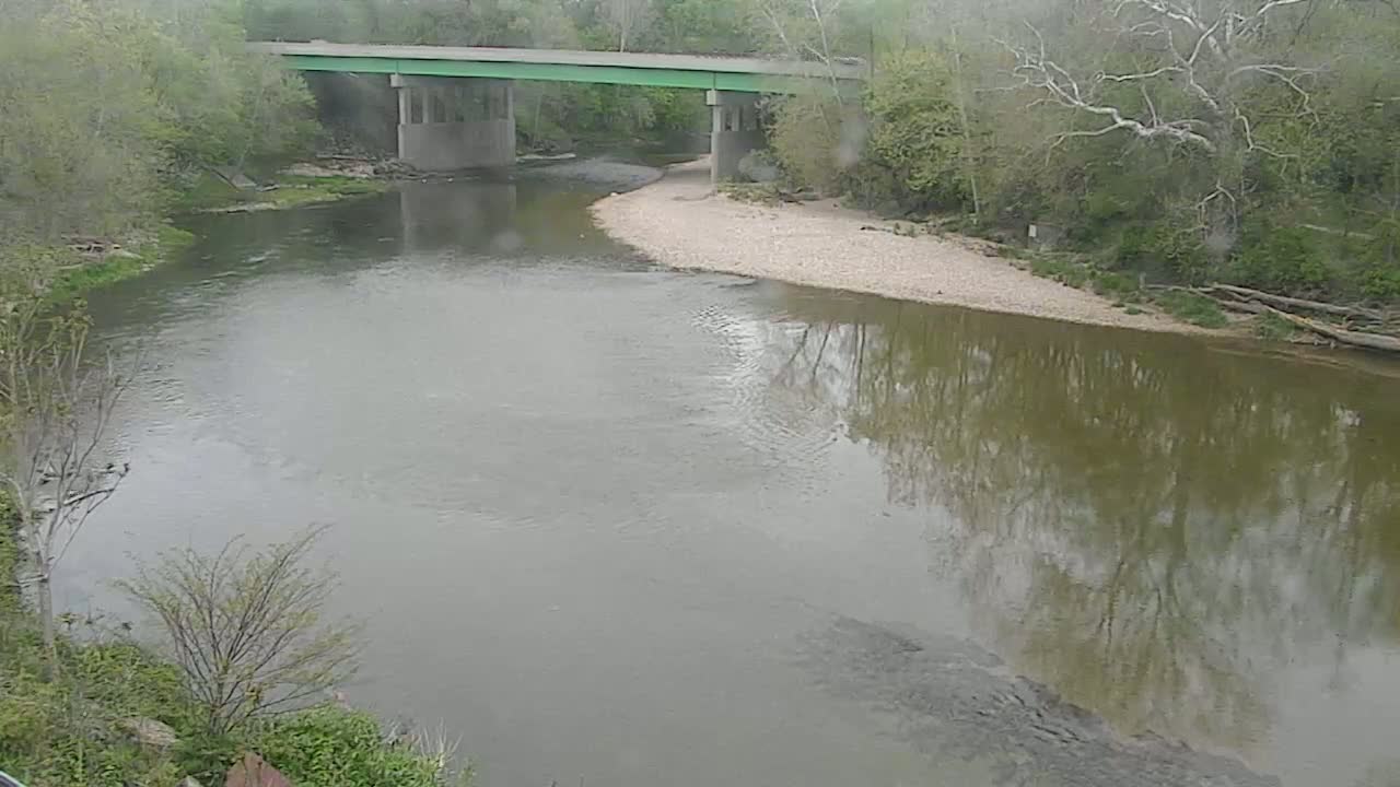 A calm, muddy river flows under a concrete bridge on a cloudy day, bordered by lush green trees and a sandy riverbank.