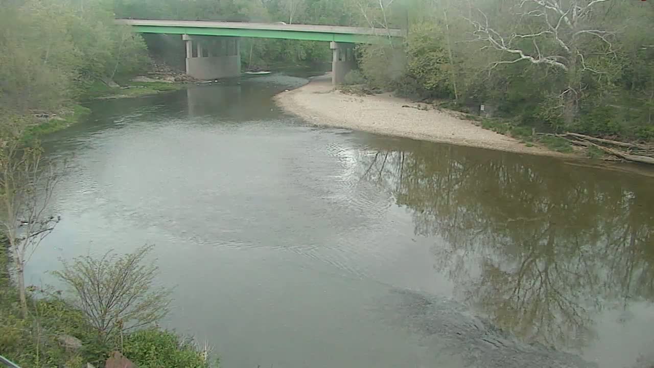 A river flows under a bridge on an overcast day, bordered by trees and a sandy bank.