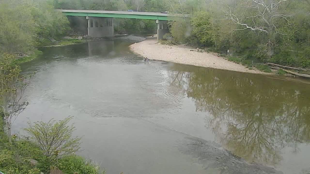 A river flows under a bridge on an overcast day, with a few people wading in the shallows near the bank.
