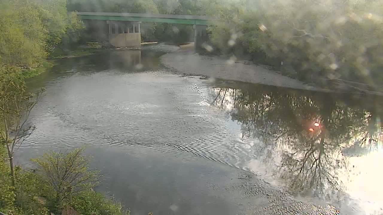 A river flows under a bridge on a slightly overcast day, with the reflection of trees and sky visible on the water's surface.