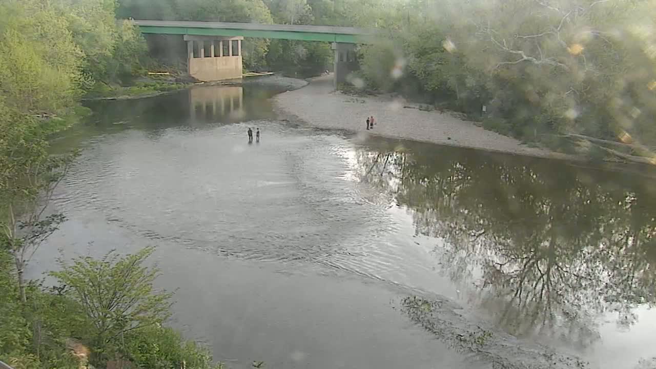 A river flows under a bridge on an overcast day, with a few people standing on the riverbank.