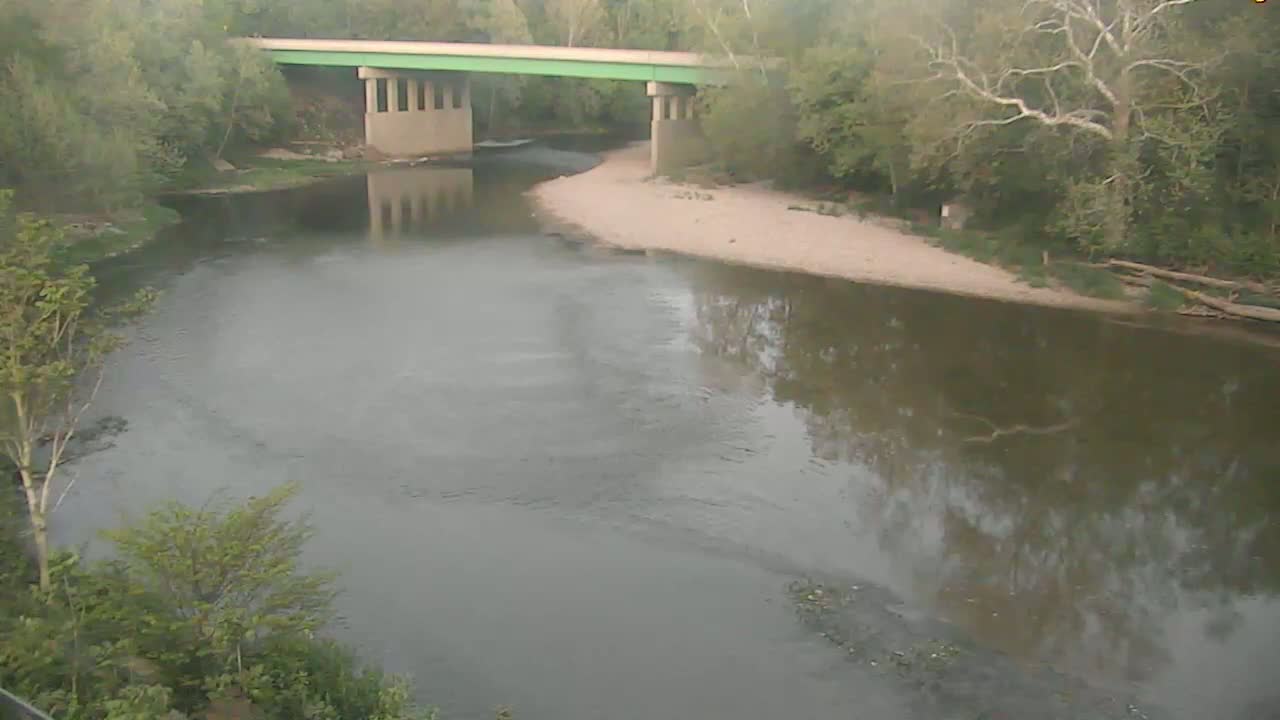 A calm river reflects the overcast sky and surrounding trees, with a bridge spanning the waterway in the background.