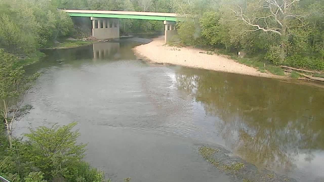 A river bends gently beneath a green bridge, surrounded by lush green trees and a sandy riverbank under a partly cloudy sky.